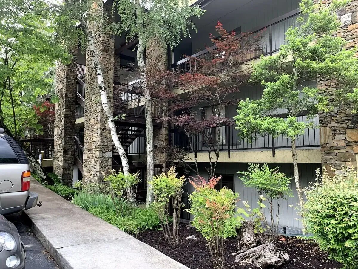 The exterior of a building with stone columns, lush greenery, a staircase, and balconies overlooking the landscape. The scene suggests peaceful strolls through shaded grounds and time spent enjoying the outdoors together.