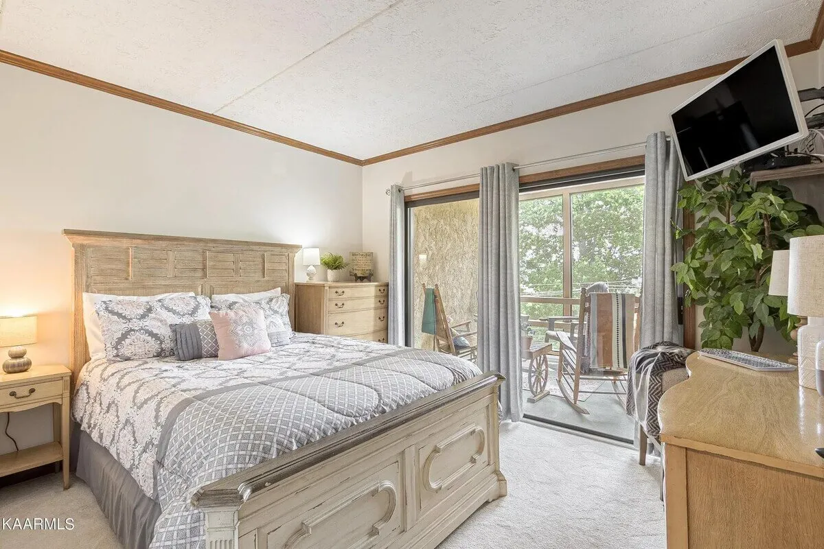 A cozy guest bedroom with a bed adorned with decorative pillows, a large window offering views of the outdoors, a wooden dresser, and a potted green plant. Natural light fills the space, enhancing the sense of calm and serenity.