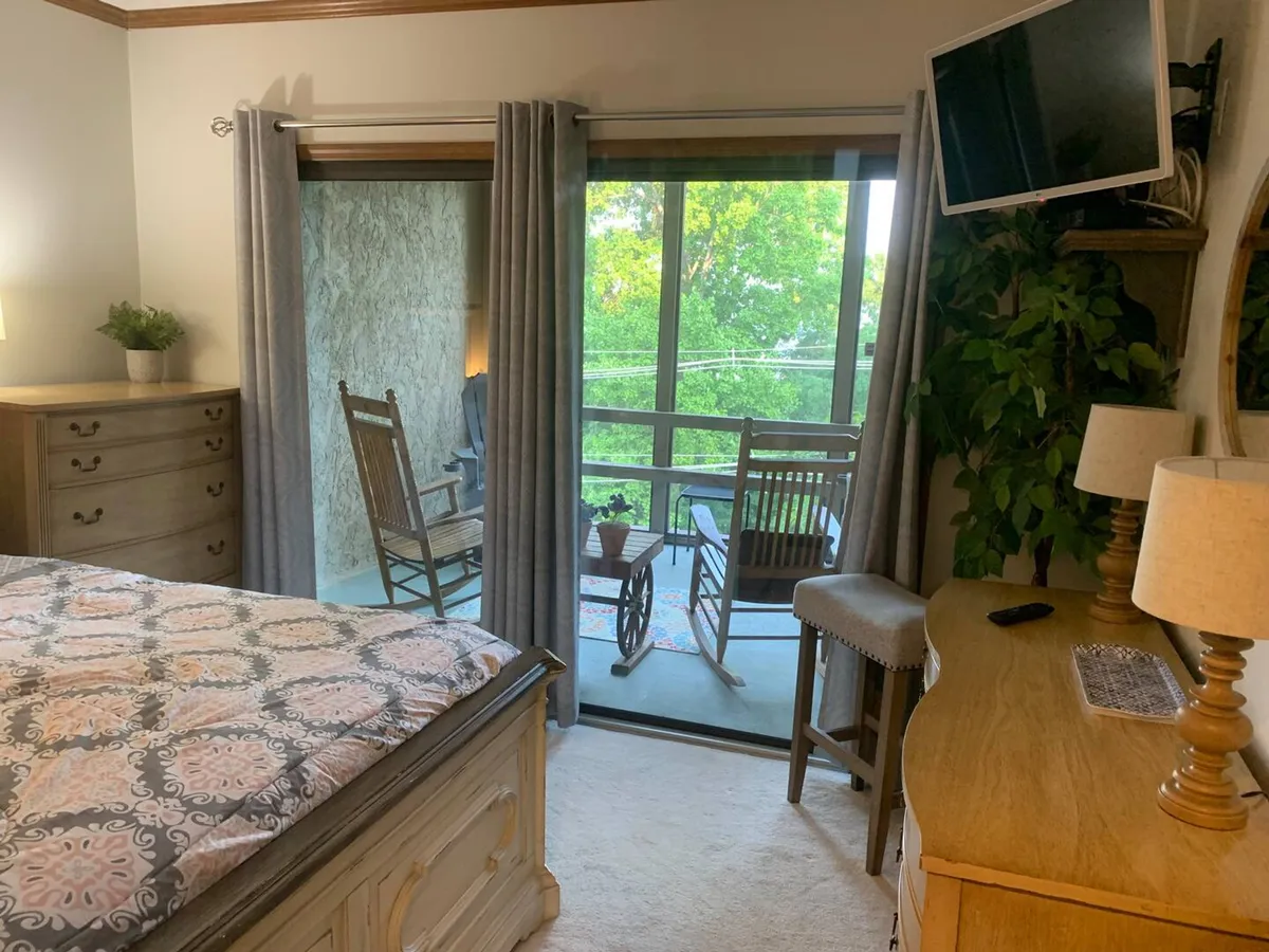 A peaceful bedroom with a bed facing a sliding glass door that opens to a balcony with rocking chairs. A dresser and potted plant complement the room, while the balcony invites serene moments with mountain or treetop vistas.