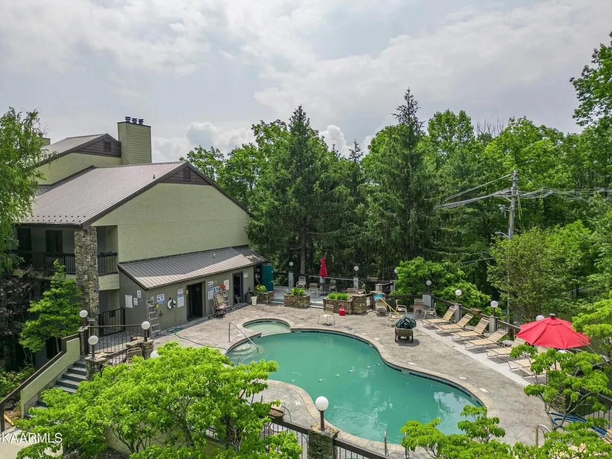 A sunny outdoor swimming pool area surrounded by lush greenery and lounge chairs, with a red umbrella adding a pop of color and a view of the condo building facade in the background. The setting is relaxed and inviting, perfect for swimming, sunbathing, or family gatherings after a day outdoors.