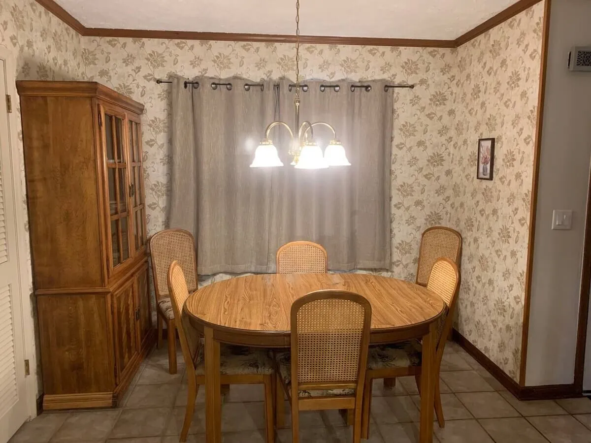 A formal dining area featuring a sturdy wooden dining table, a central chandelier, elegant floral wallpaper, and window drapes. The setting exudes warmth, making it perfect for family dinners, games, or celebrating special occasions together.