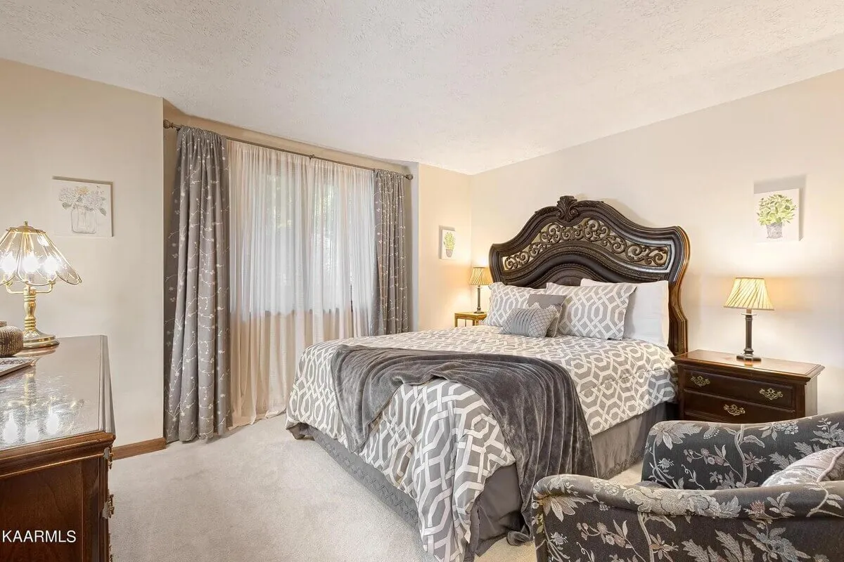 A serene master bedroom featuring an ornate bed frame with decorative pillows, a plush armchair by the window, an elegant lamp for ambient lighting, and floral artwork on the wall. The plush bedding and cozy layout evoke a restful, upscale retreat.