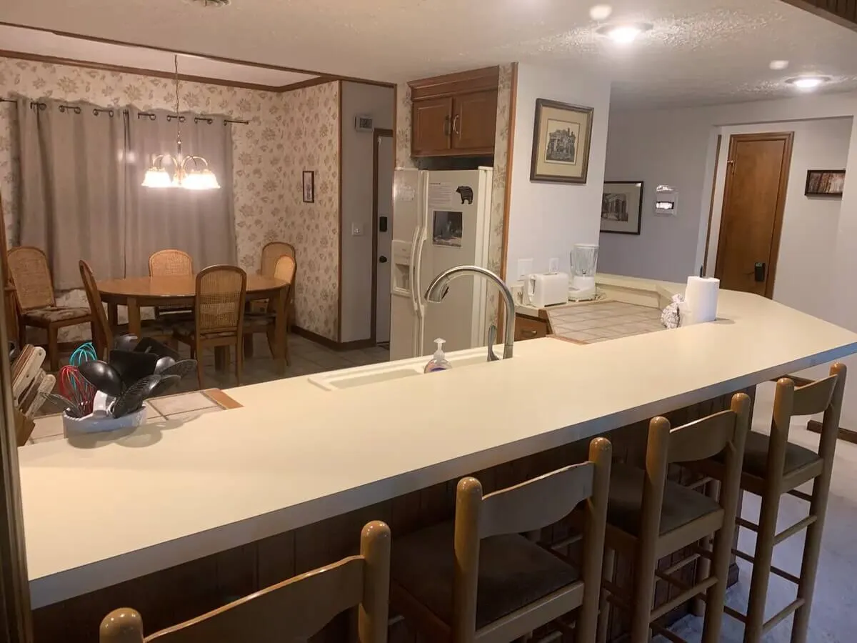 A wide view of an open-plan kitchen and dining area, featuring a kitchen island, a dining table with multiple chairs, and a refrigerator in the background. Natural lighting and soft curtains add coziness, while the layout encourages socializing and group meals.