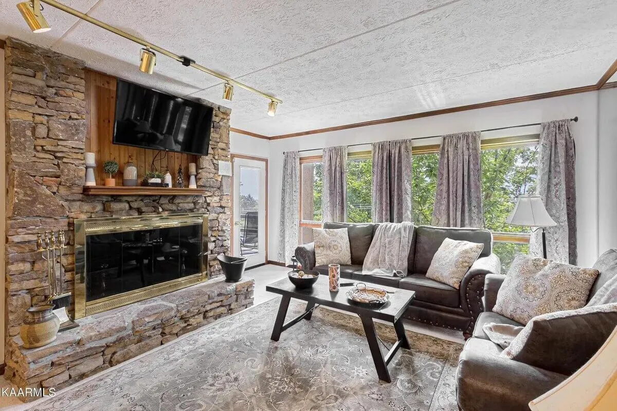 This image presents a cozy, open-concept living area featuring a stone fireplace, a comfortable sofa, a coffee table, and a television. Large windows are visible, allowing natural light to flood the space and hinting at possible scenic views beyond. The room has a welcoming atmosphere perfect for gatherings, relaxation, or sharing stories by the fire.